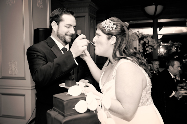black and white photo - the happy couple feeding each other wedding cake - bride is wearing white floral hairpiece -  photo by Houston based wedding photographer Adam Nyholt